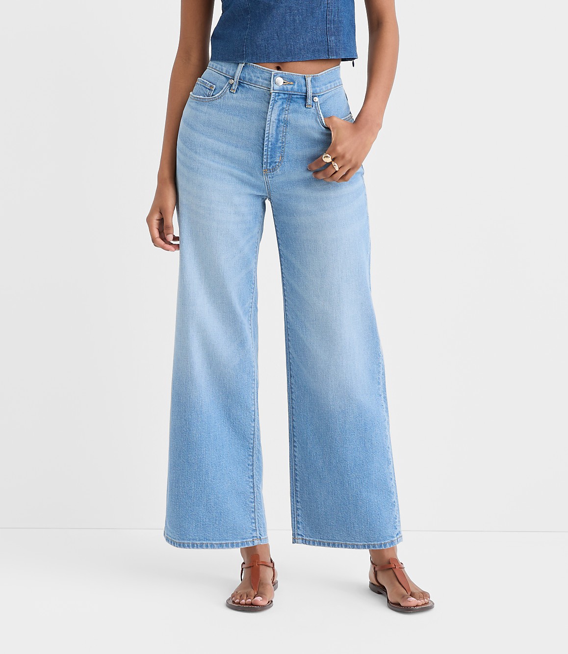 The image shows a pair of light-washed denim wide-leg jeans worn by a person, with a cropped denim top and sandals visible in the foreground against a plain white background.