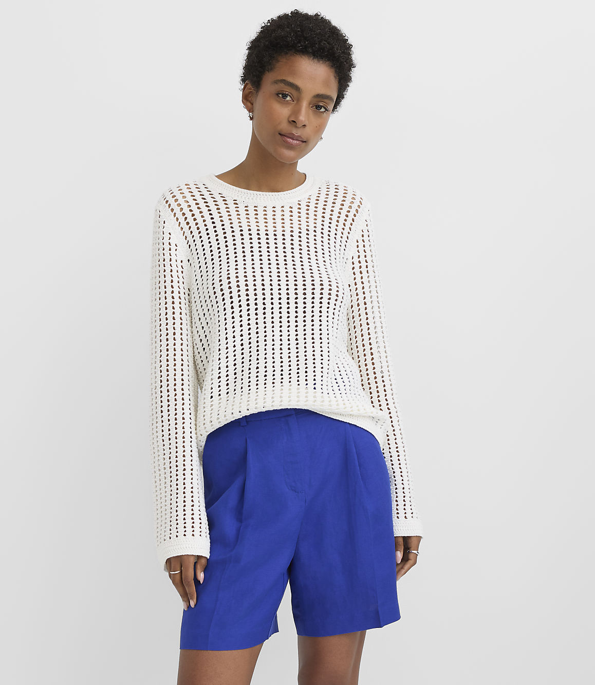 A woman in a white open-knit sweater and blue shorts stands against a plain white background.