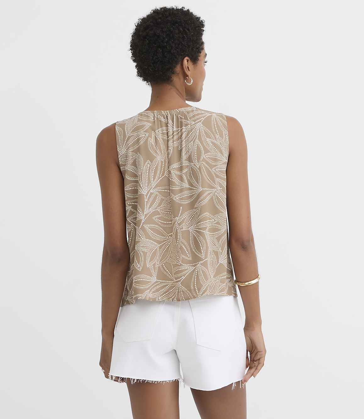 A woman in a tan, leaf-patterned sleeveless top and white denim shorts stands against a plain white background.