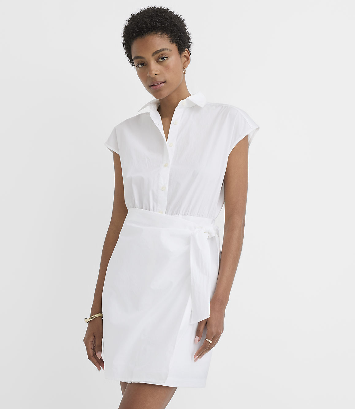 A woman in a white shirt dress with a tie waist stands against a plain white background.