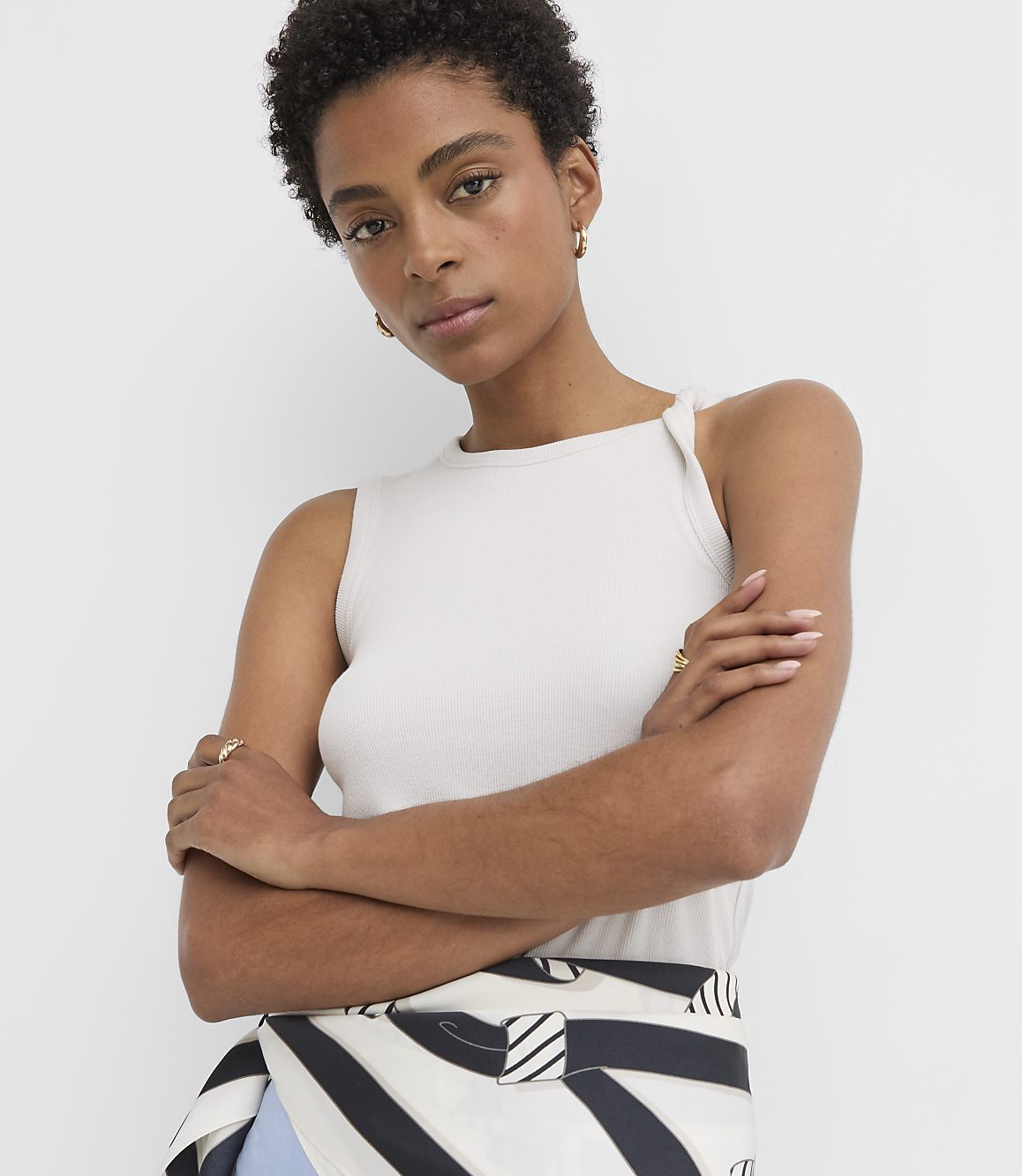 A woman with short dark hair and a light-colored tank top poses with her arms crossed in front of a plain white background.