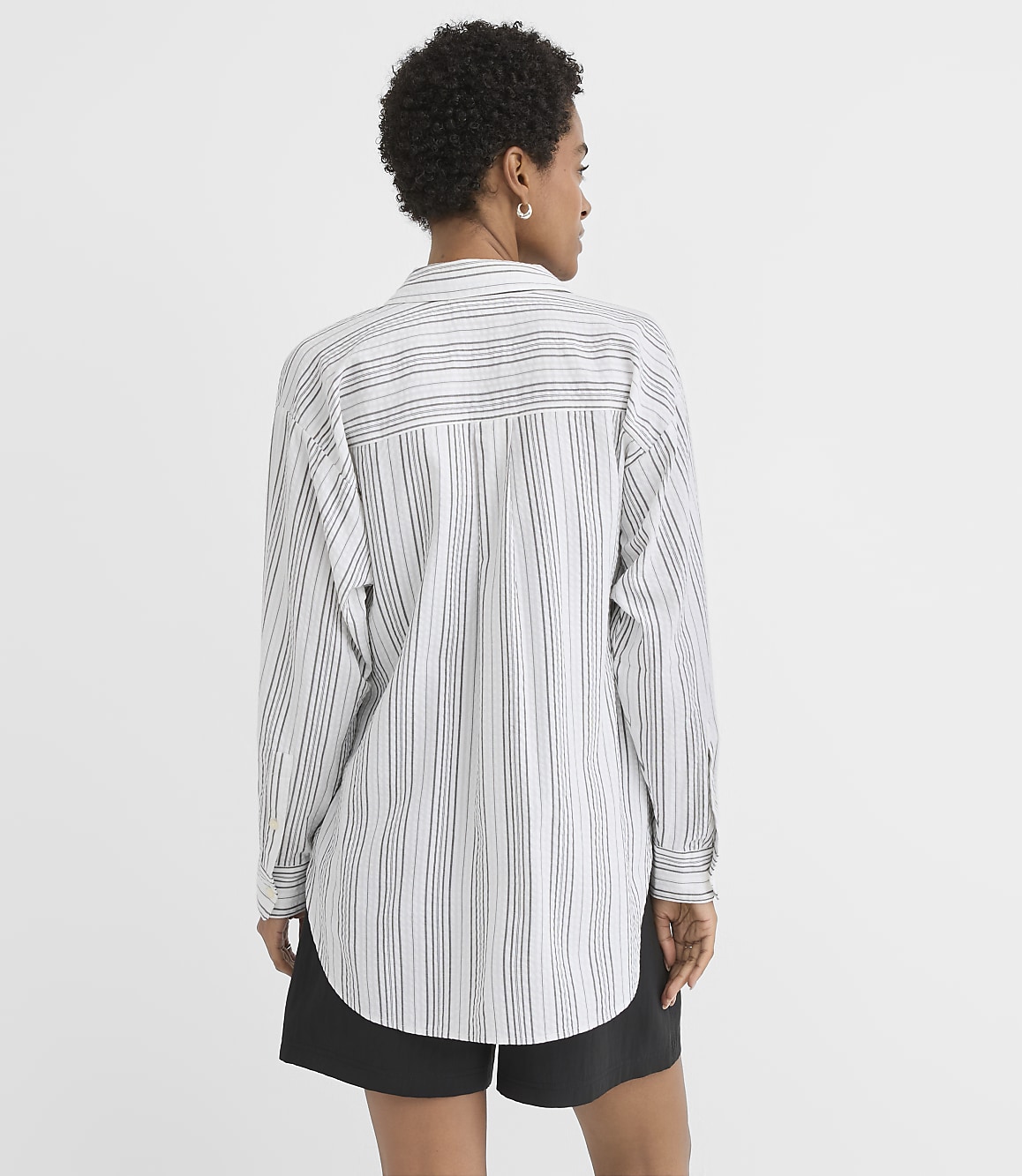 A woman from behind, wearing a striped button-down shirt and black shorts, stands against a plain white background.