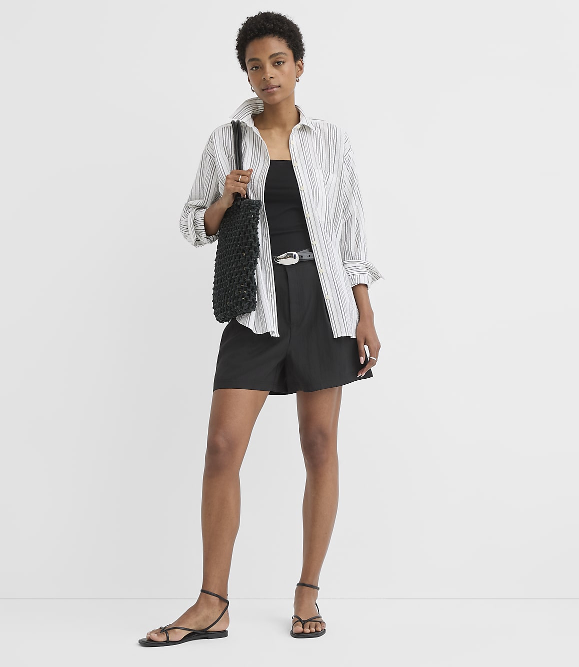 A woman in a striped shirt and black shorts stands against a plain white background, holding a black woven bag.