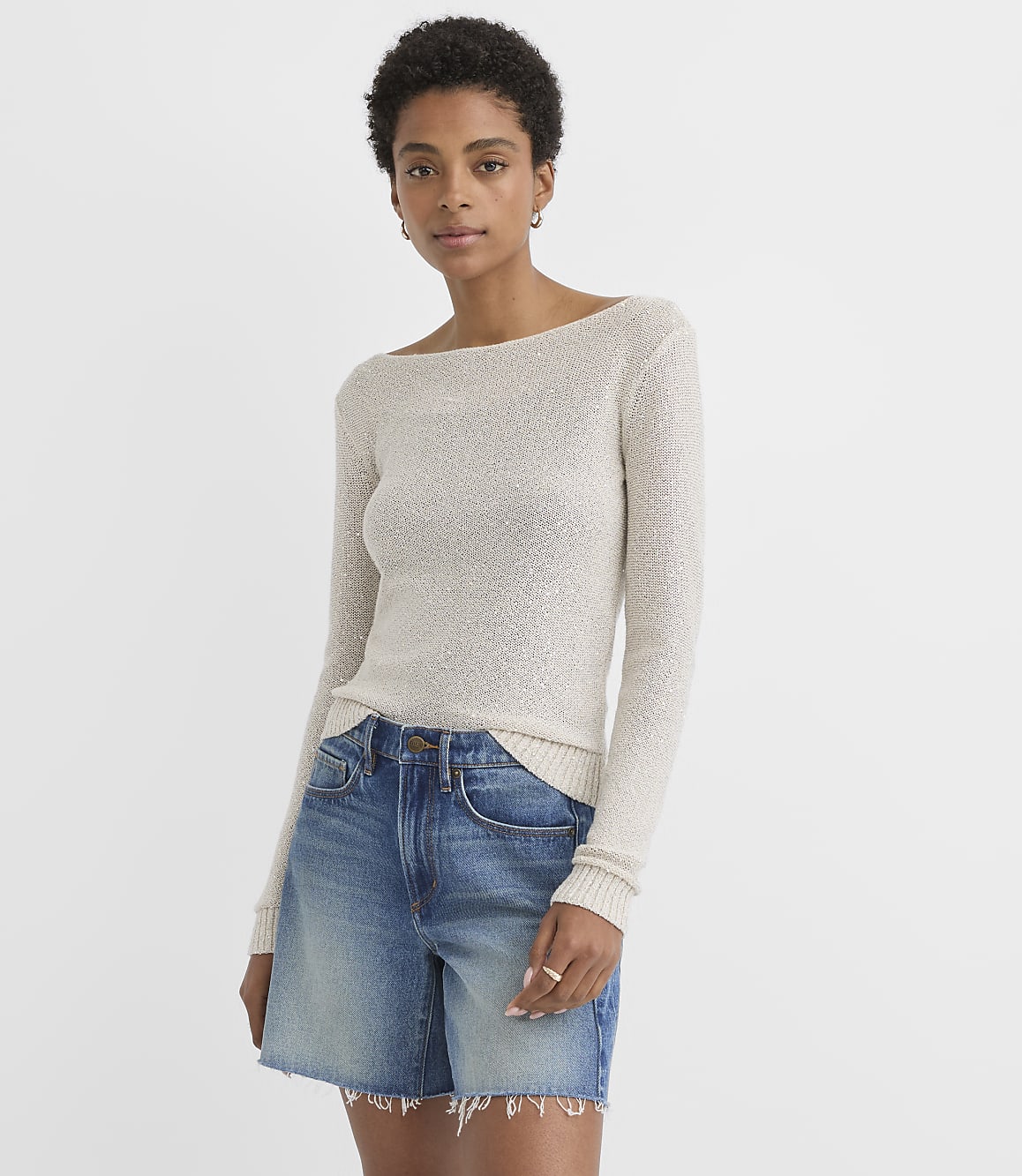 A woman in a cream-colored boatneck sweater and denim shorts stands against a plain white background.