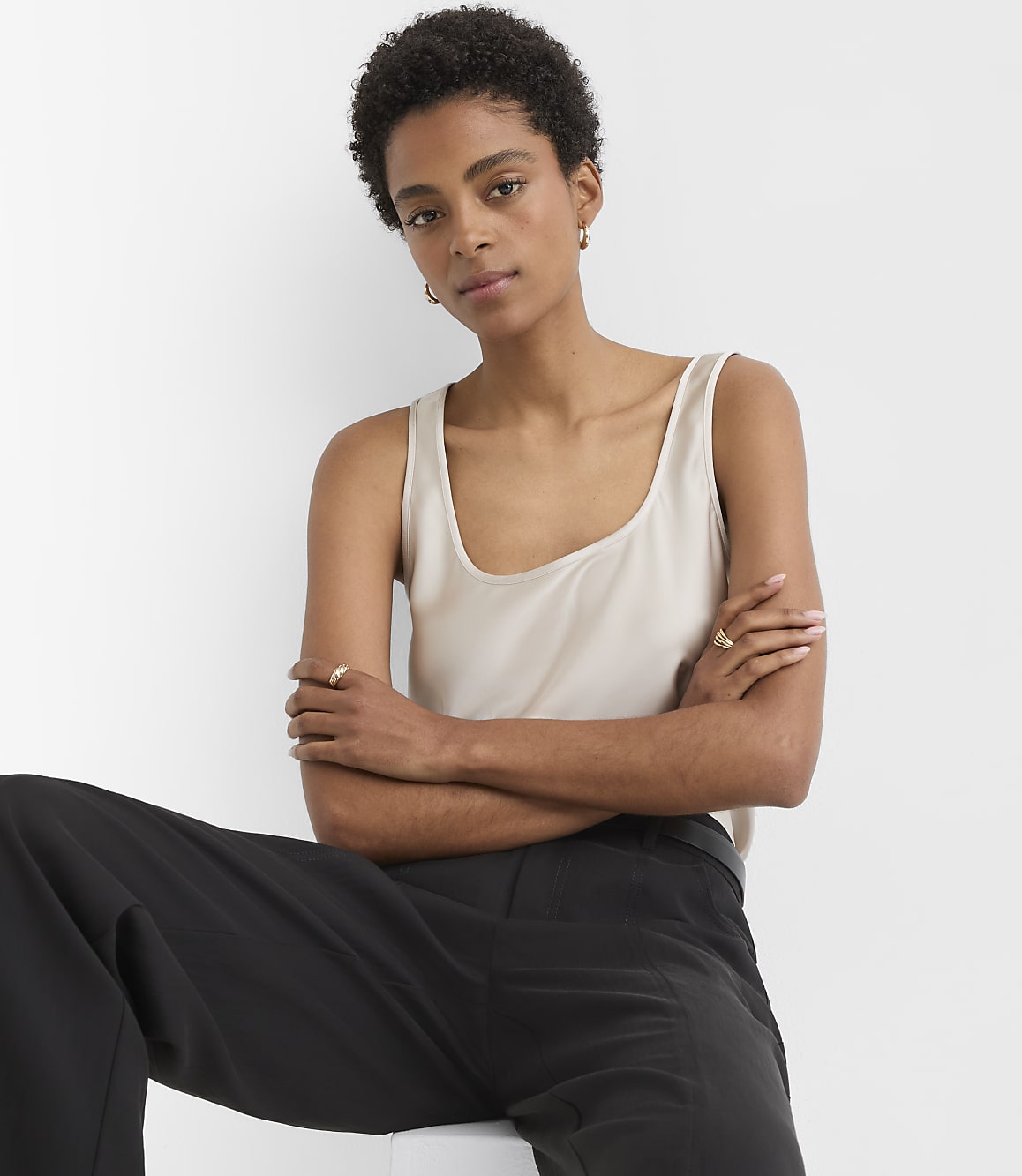 A woman in a beige tank top and black pants sits with her arms crossed against a plain white background.