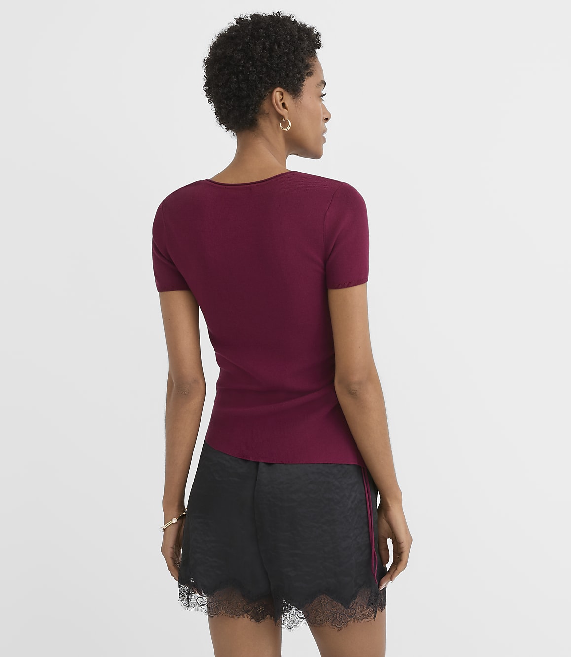 A woman in a maroon t-shirt and black lace-trimmed shorts stands against a plain white background.