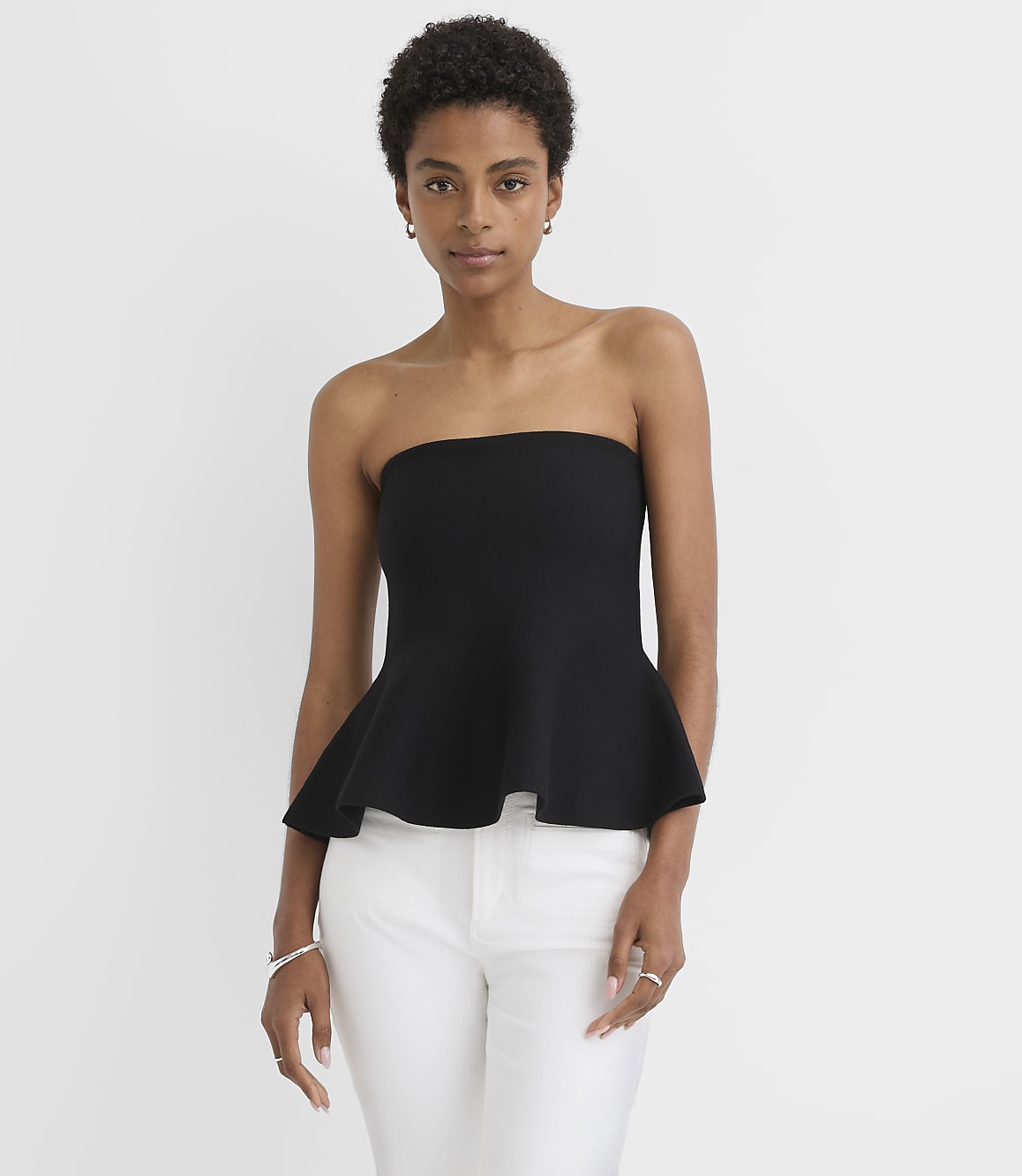 A woman in a black strapless peplum top and white pants stands against a plain white background.