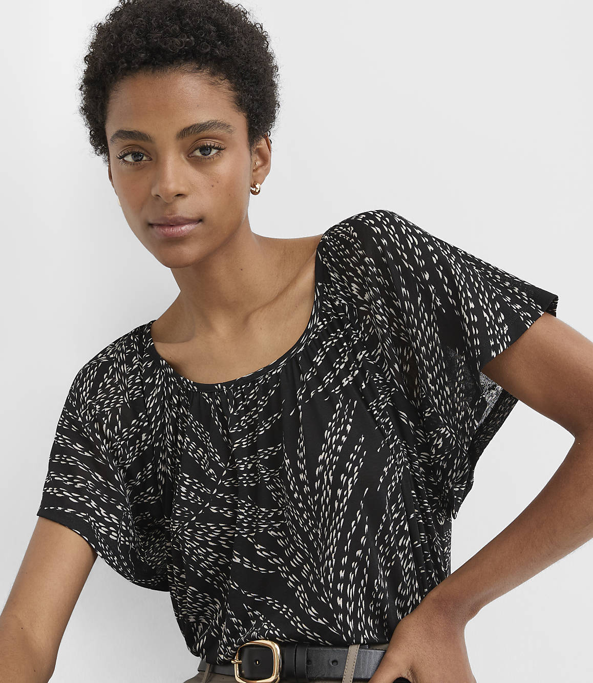 A woman with short dark hair and a black and white patterned top is shown against a plain white background.