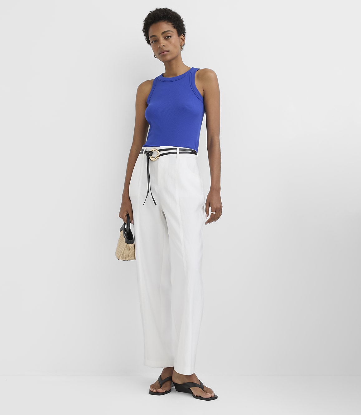 A woman in a blue tank top and white pants stands against a plain white background, holding a small straw bag.