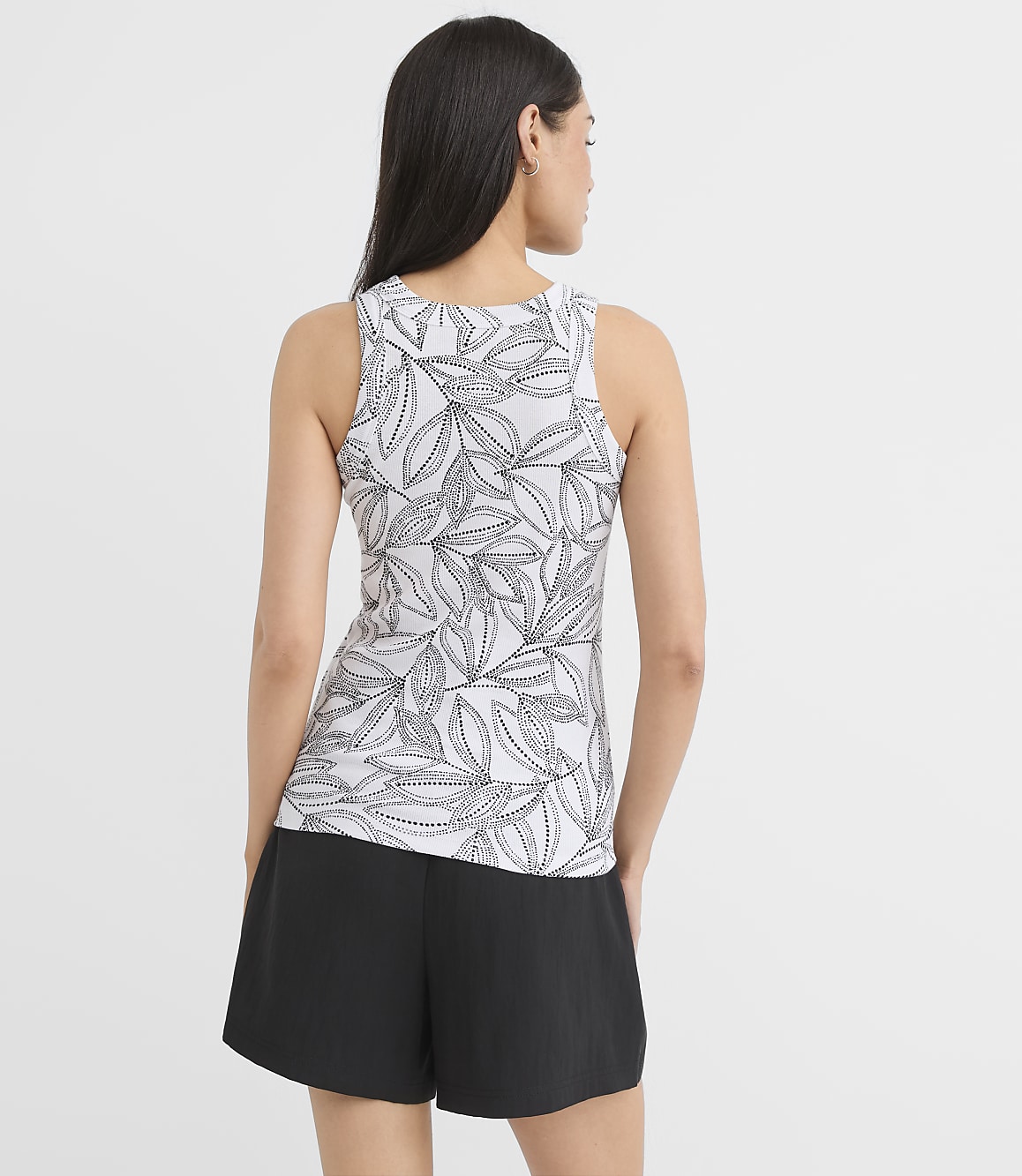 A woman in a leaf-patterned tank top and black shorts stands against a plain white background.