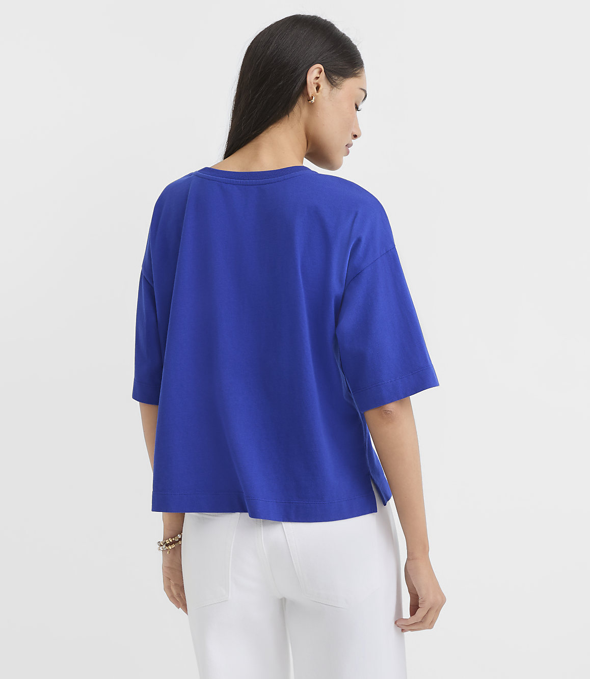 A woman in a blue t-shirt and white pants stands against a plain white background.
