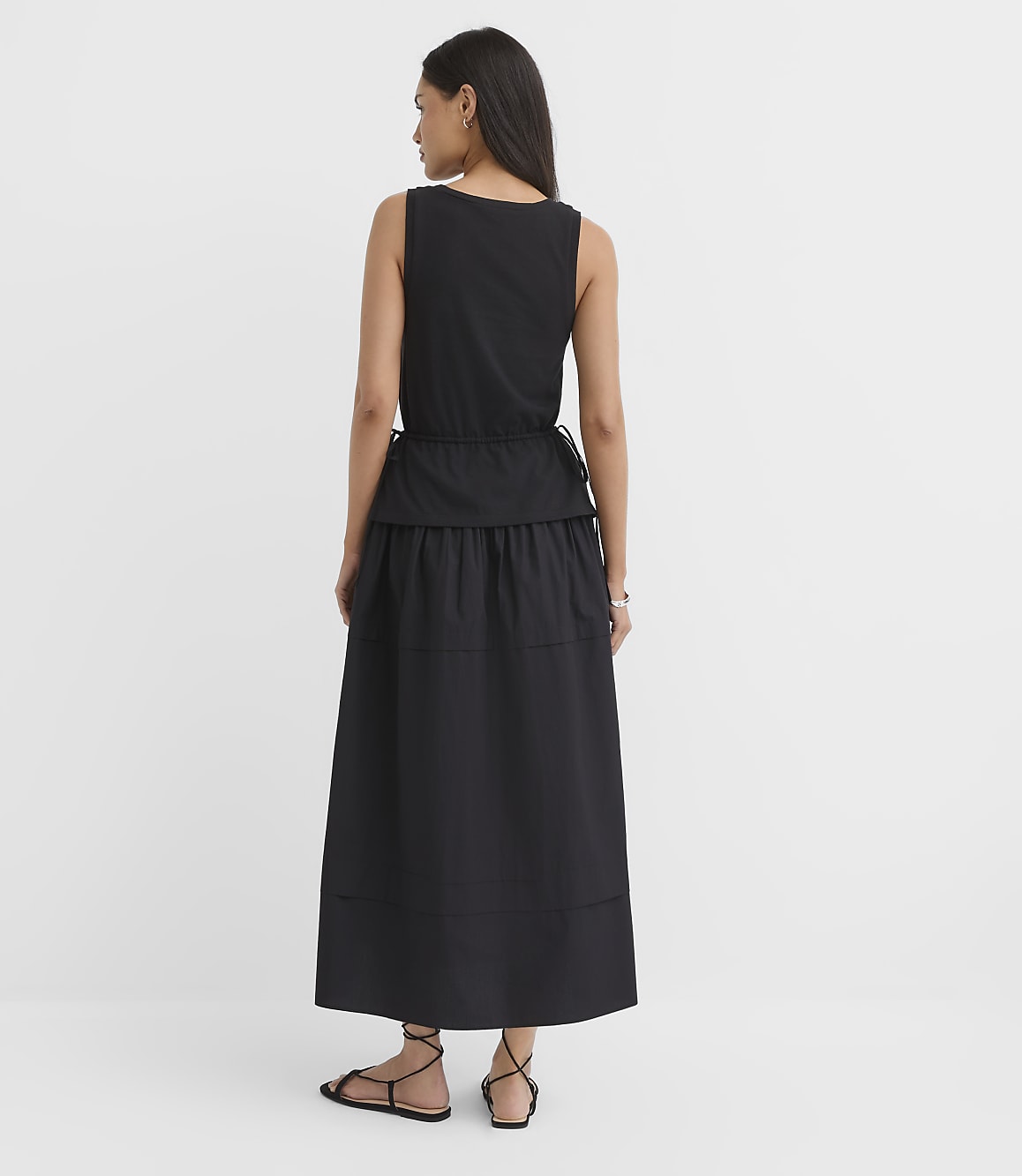 A woman in a black tiered dress and sandals stands against a plain white background.