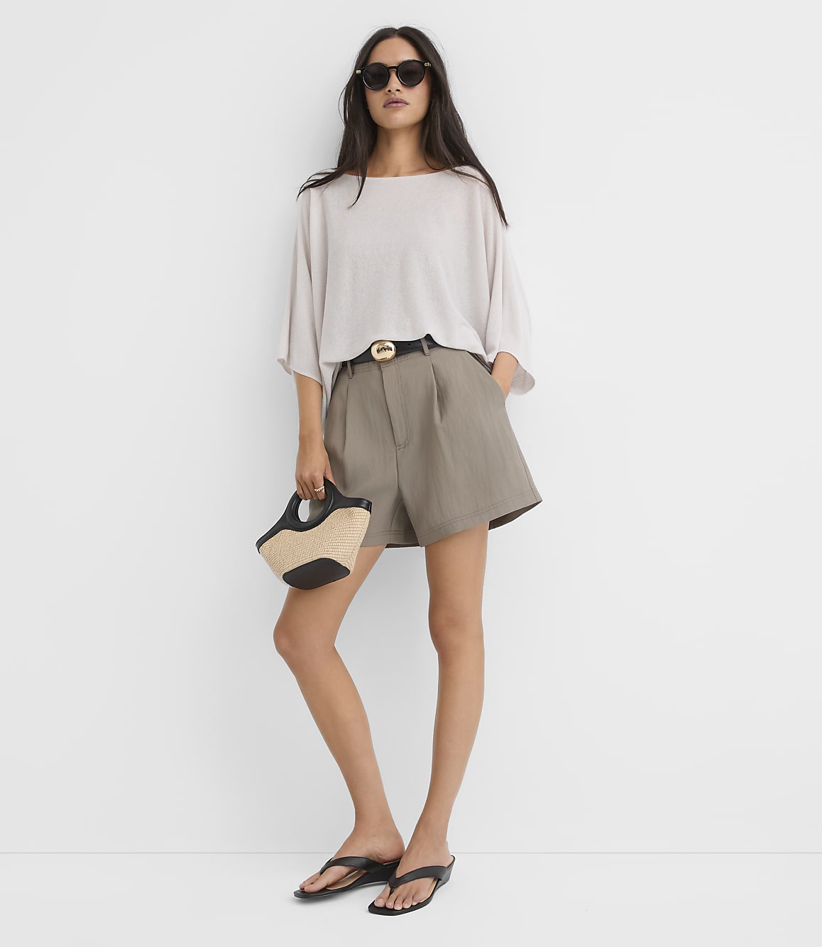 A woman in a light-colored oversized top, taupe shorts, and black sandals stands against a plain white background, holding a straw and black handbag.