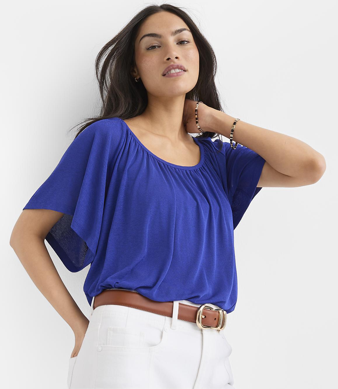 A woman in a blue top and white pants stands against a plain white background.
