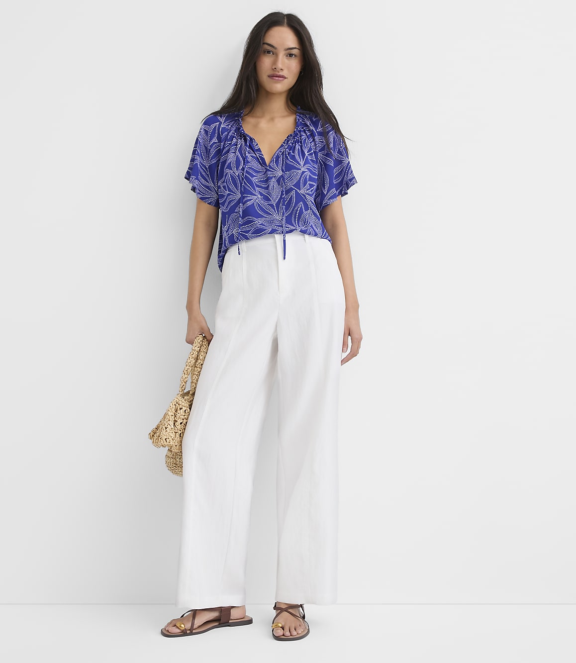 A woman in a blue floral top and white pants stands against a plain white background, holding a straw bag.