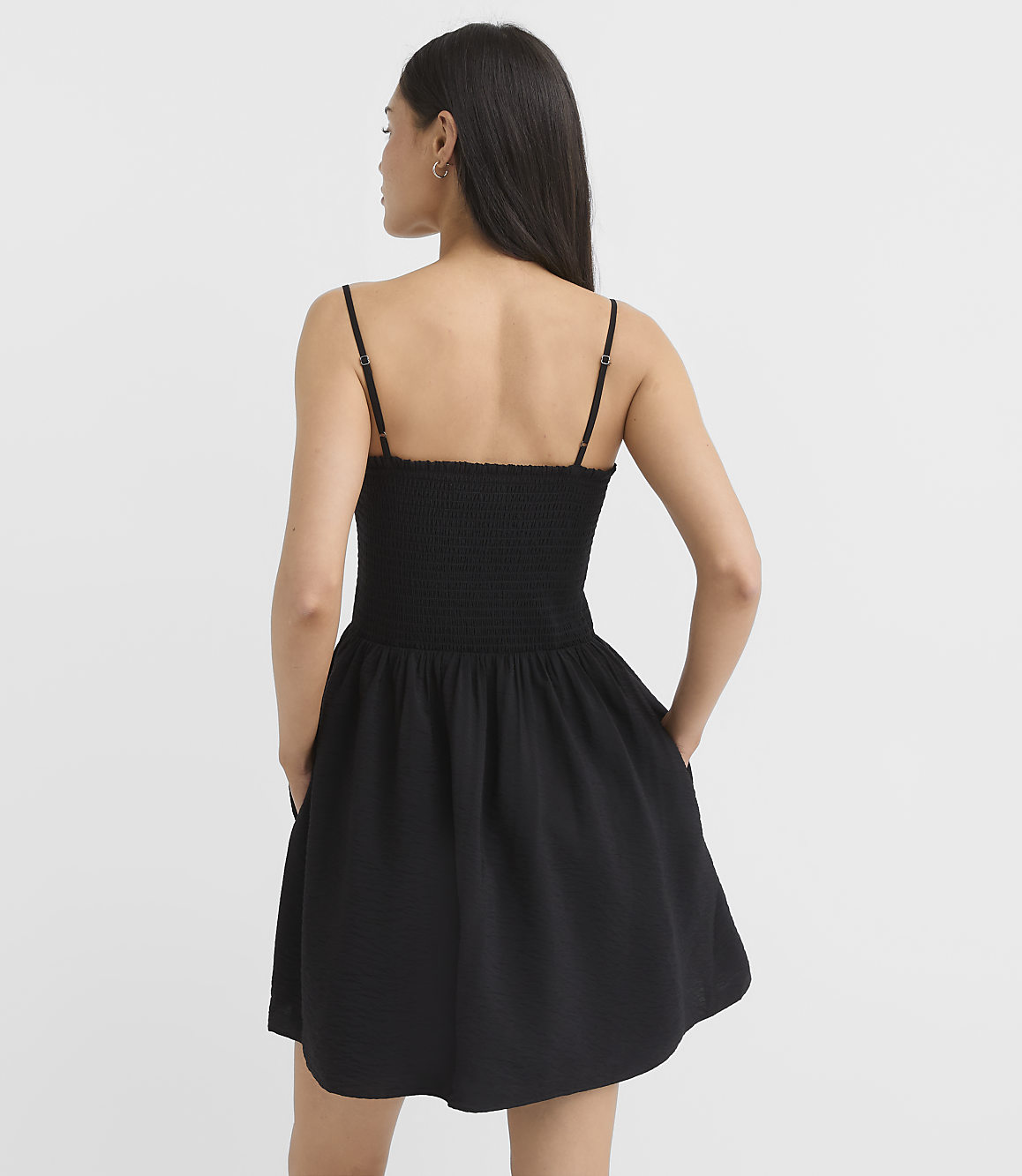 A woman in a black smocked-bodice mini dress with her hands in her pockets stands against a plain white background.