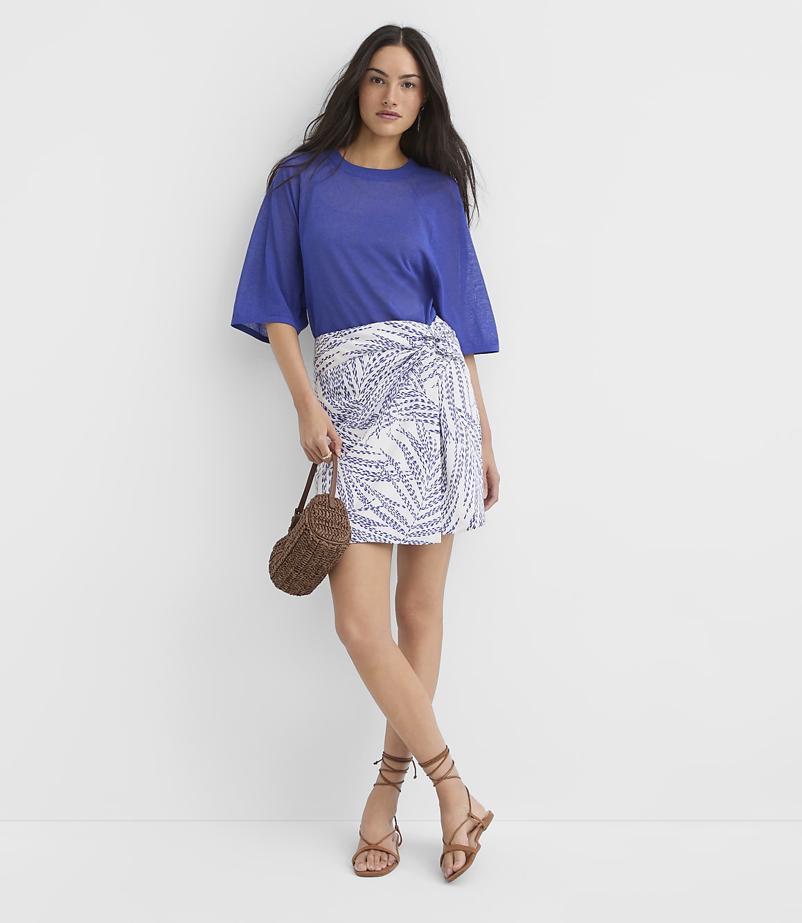 A woman in a blue top and a patterned wrap skirt stands against a plain white background, holding a woven bag.