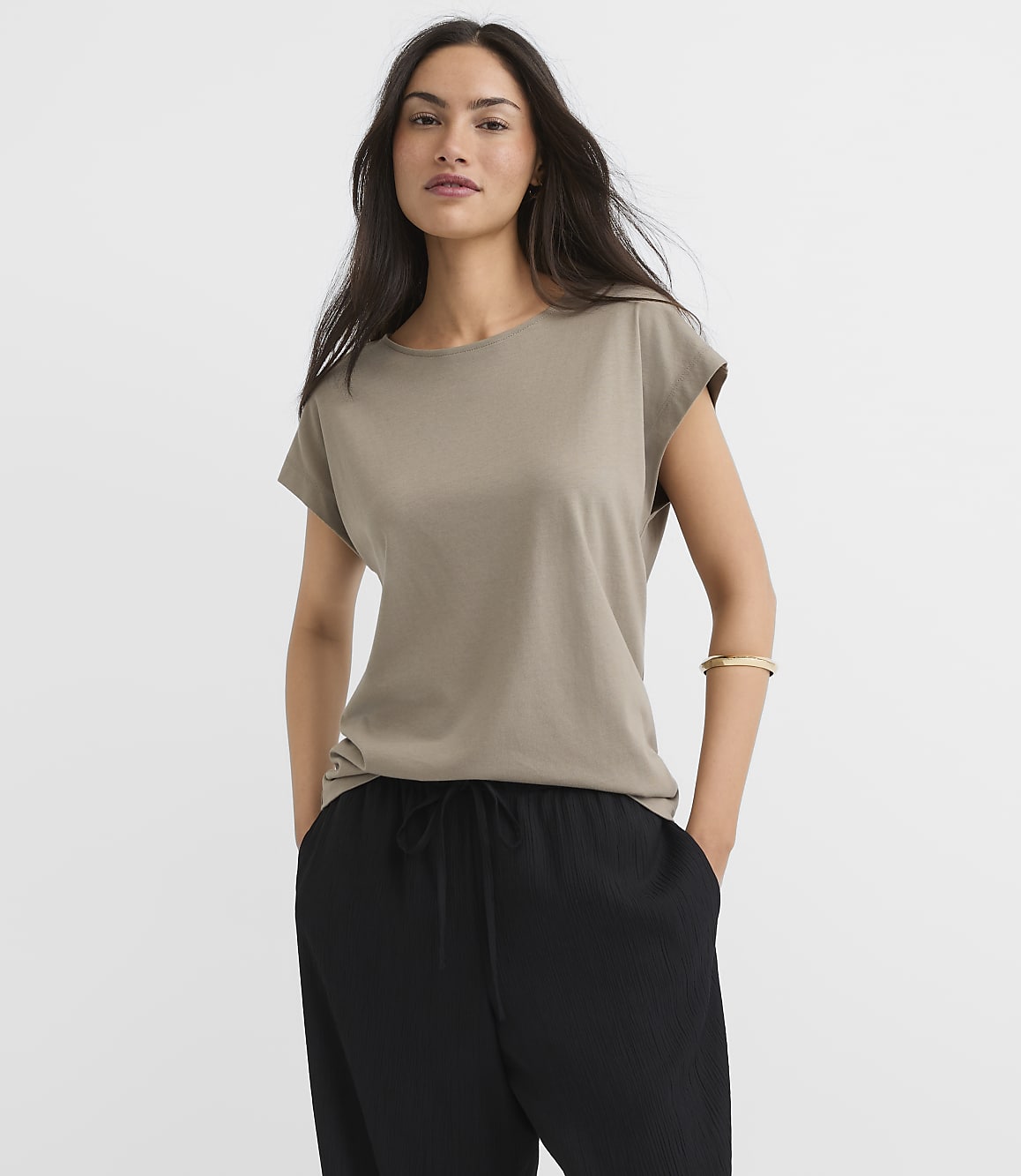A woman in a taupe t-shirt and black pants stands against a plain gray background.