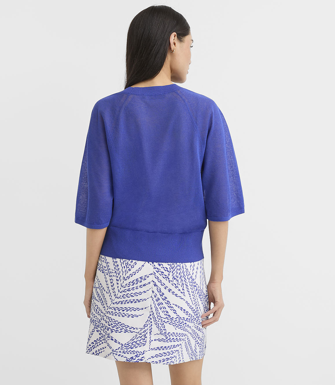 A woman in a blue sweater and patterned skirt stands against a plain white background.