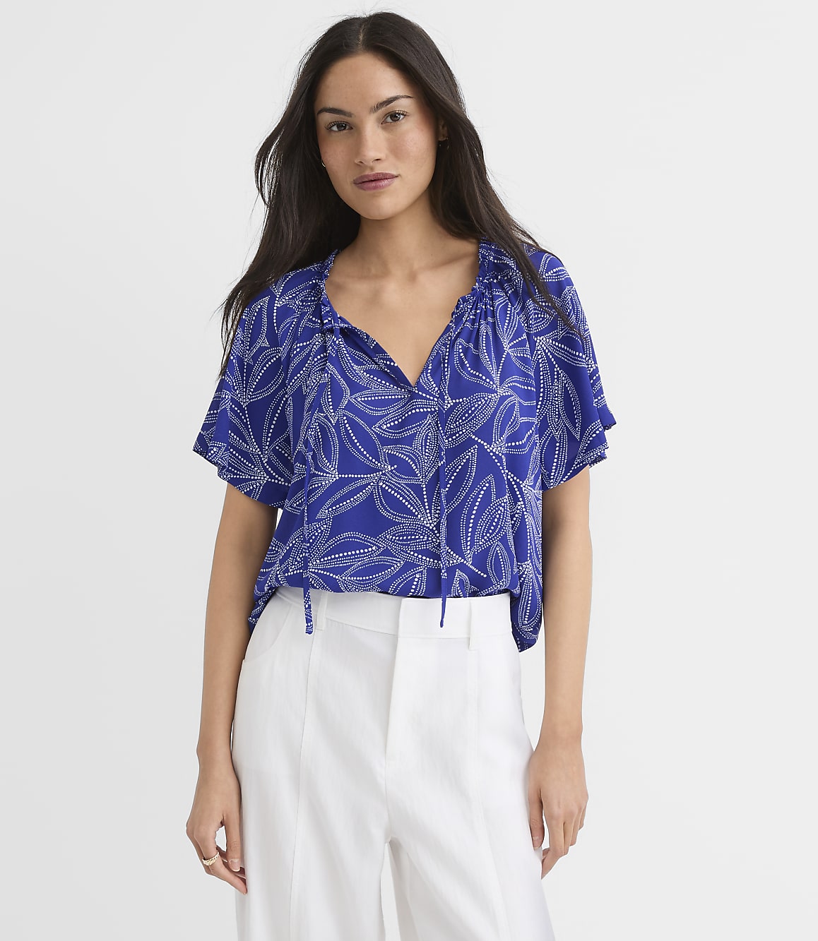 A woman in a blue floral blouse and white pants stands against a plain white background.