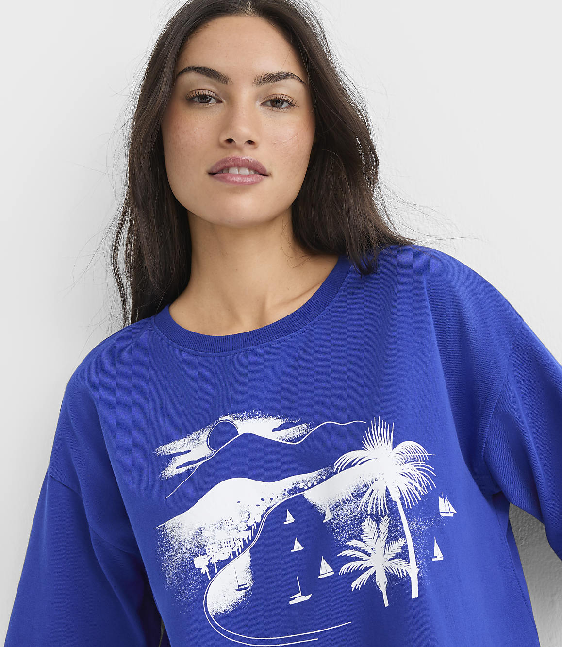 A woman in a blue sweatshirt with a white graphic of a tropical beach scene featuring palm trees and sailboats in the background.
