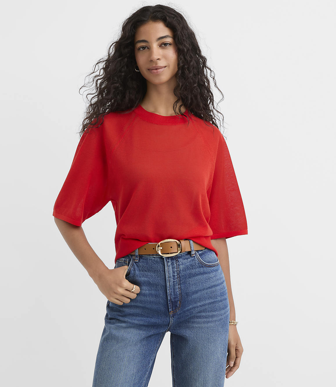 A woman in a red t-shirt and blue jeans stands against a plain white background.