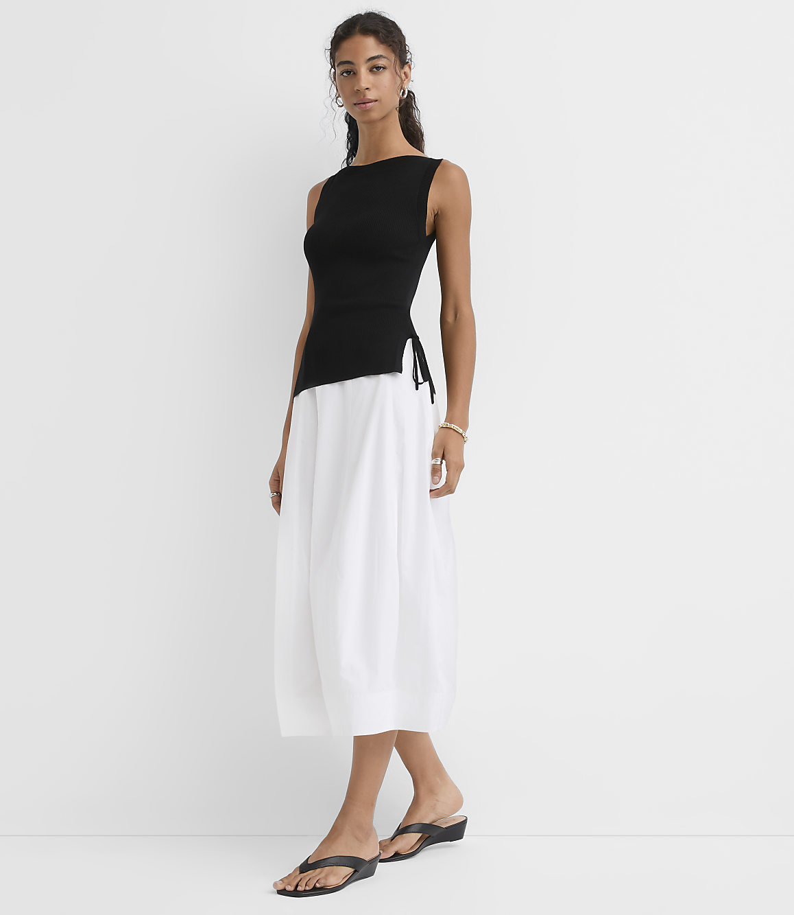 A woman in a black tank top and white skirt stands against a plain white background.
