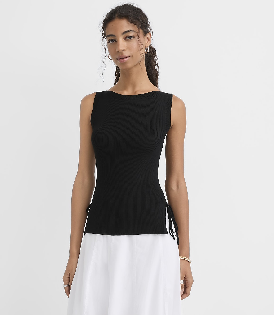 A woman in a black sleeveless top with white skirt stands against a plain white background.