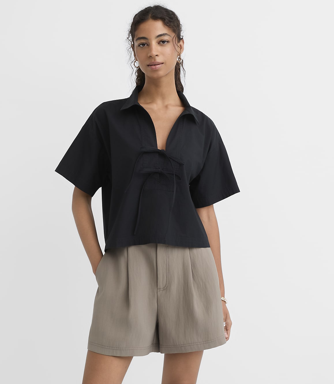 A woman in a black collared shirt and taupe shorts stands against a plain white background.