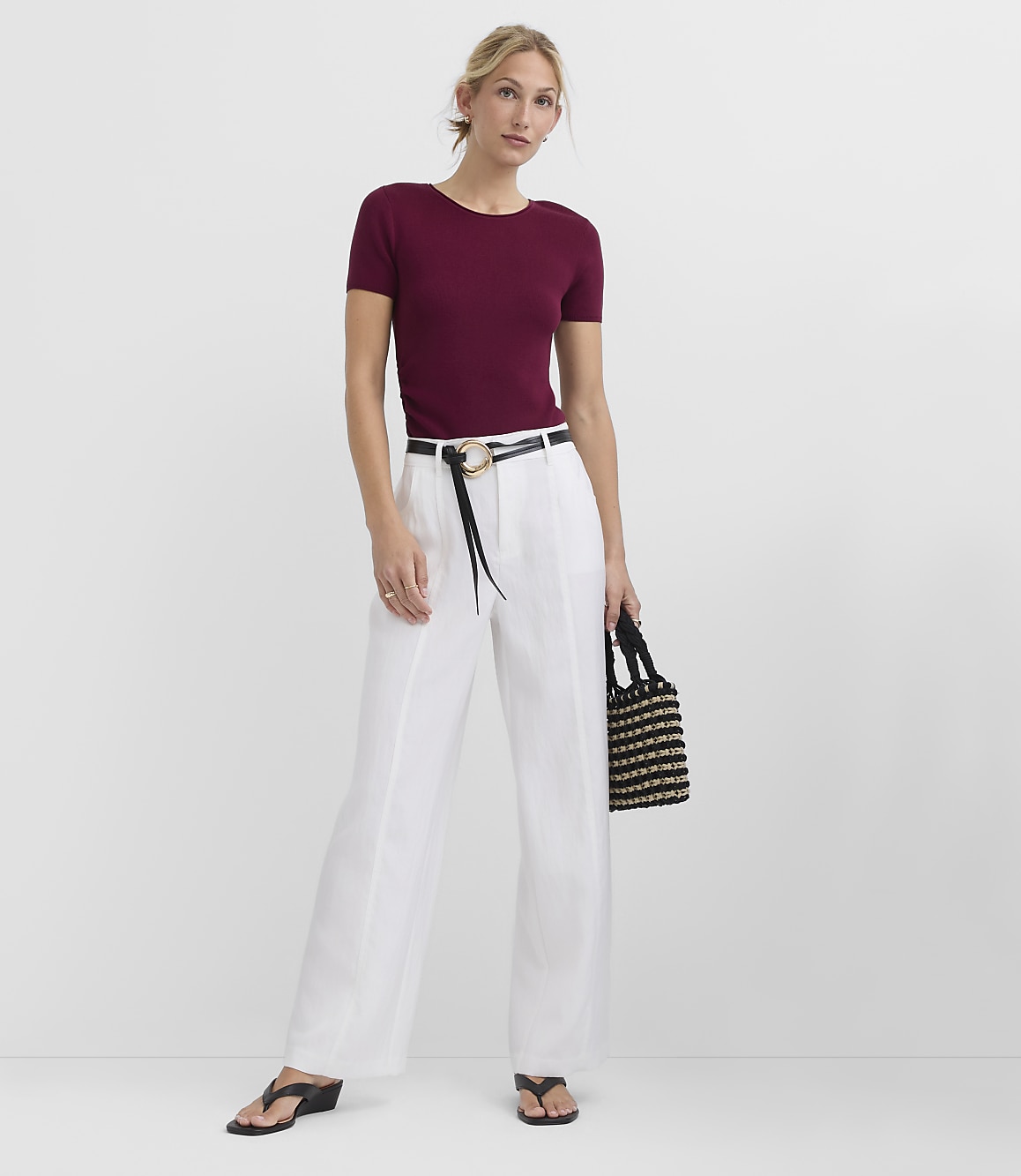 A woman in a maroon top and white pants stands against a plain gray background, holding a black and gold woven bag.