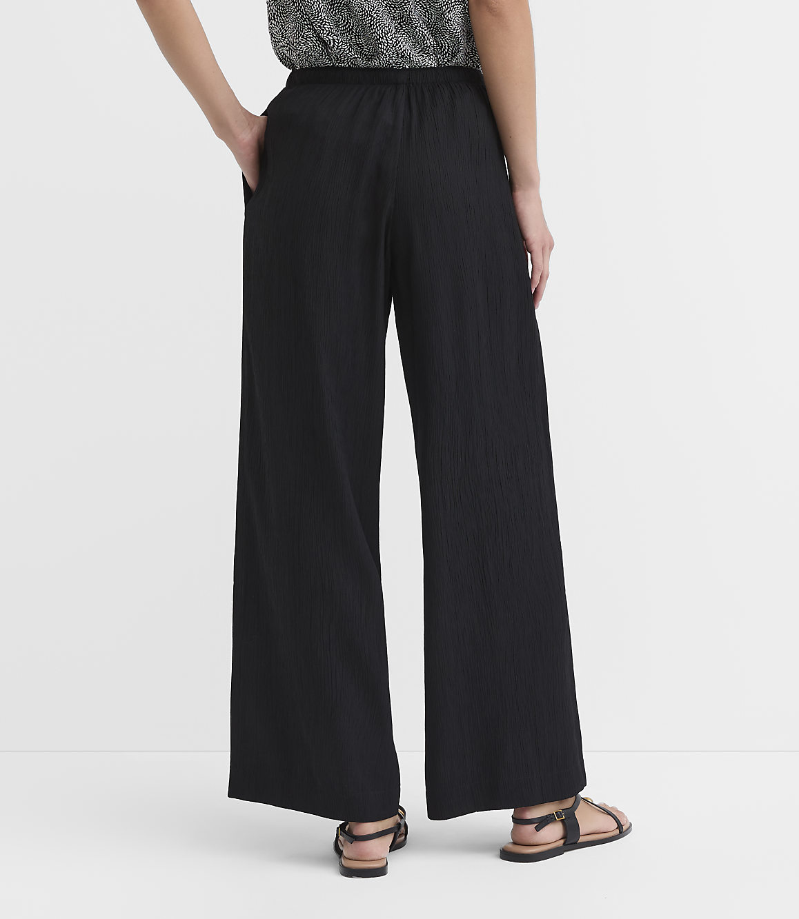 A person wearing black wide-leg pants and sandals stands against a plain white background.
