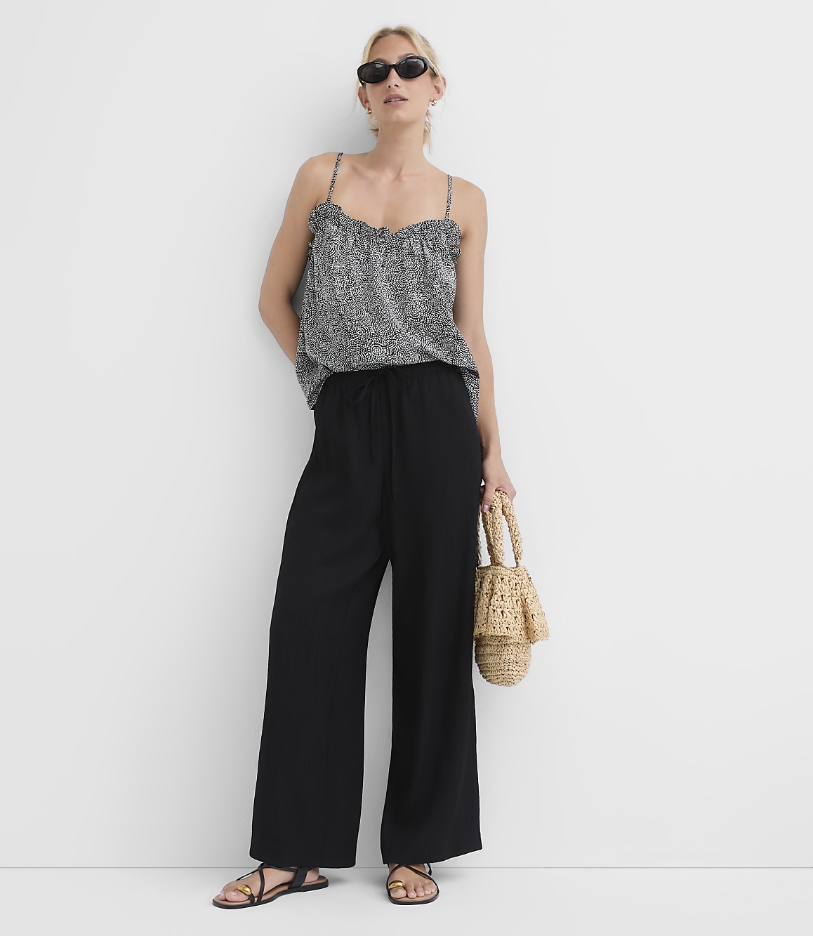 A woman in a patterned camisole and black wide-leg pants stands against a plain white background, holding a straw bag.