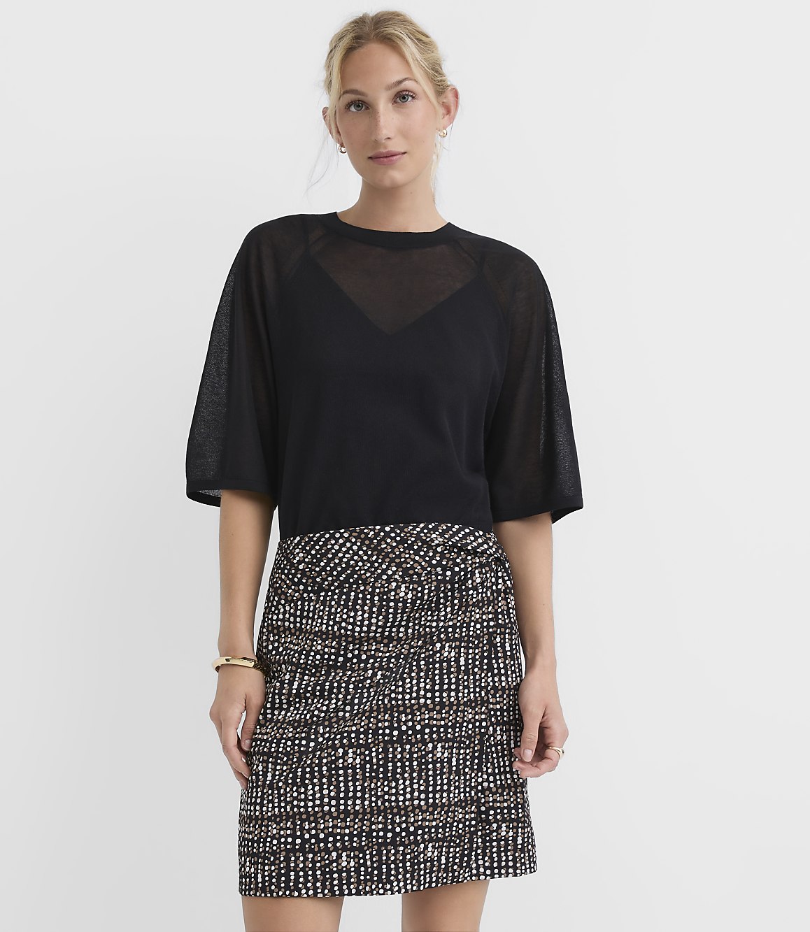 A woman in a black sheer top and a patterned skirt stands against a plain white background.