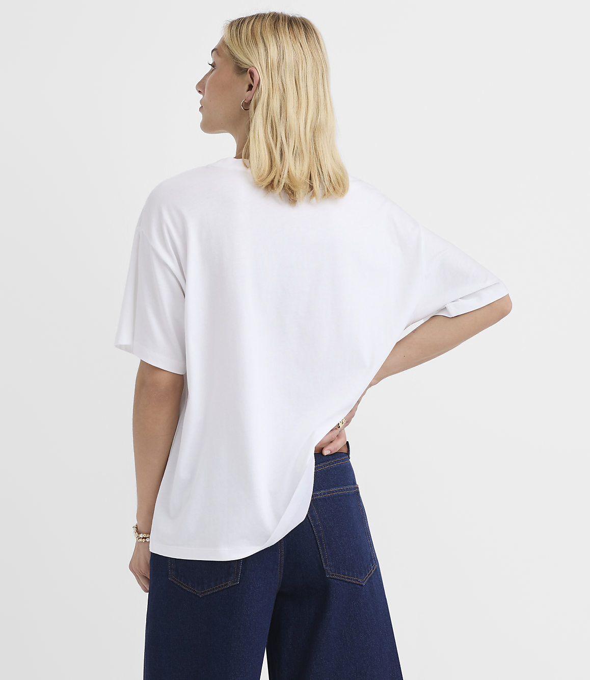 A woman in a white t-shirt and dark jeans stands against a plain white background.
