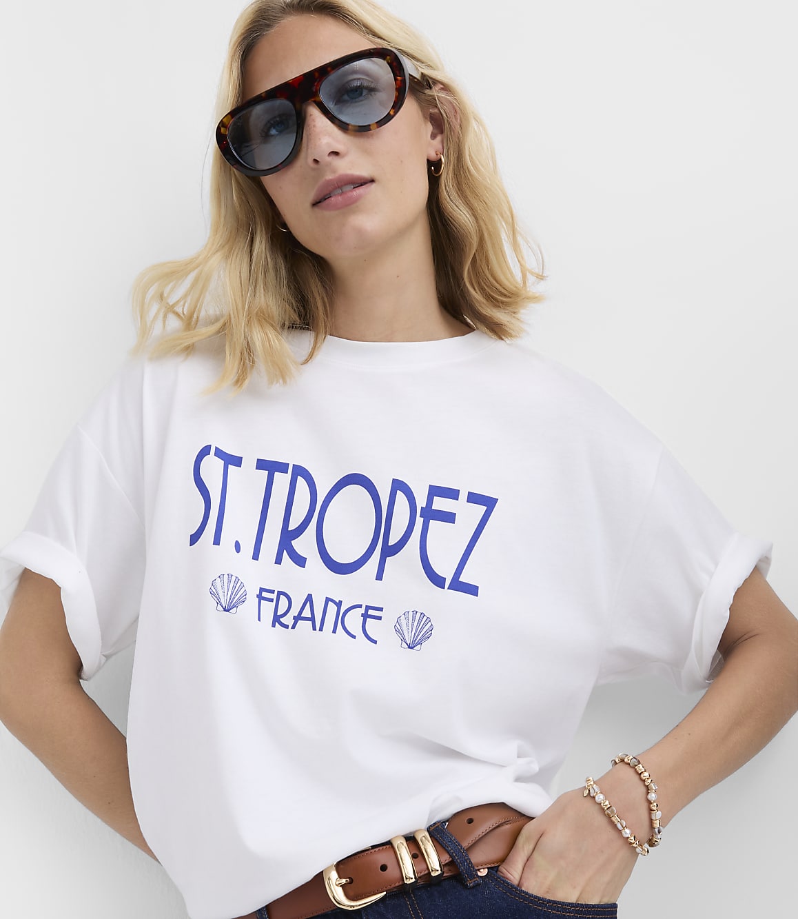 A woman in a white t-shirt with "ST. TROPEZ FRANCE" printed on it poses against a plain white background.