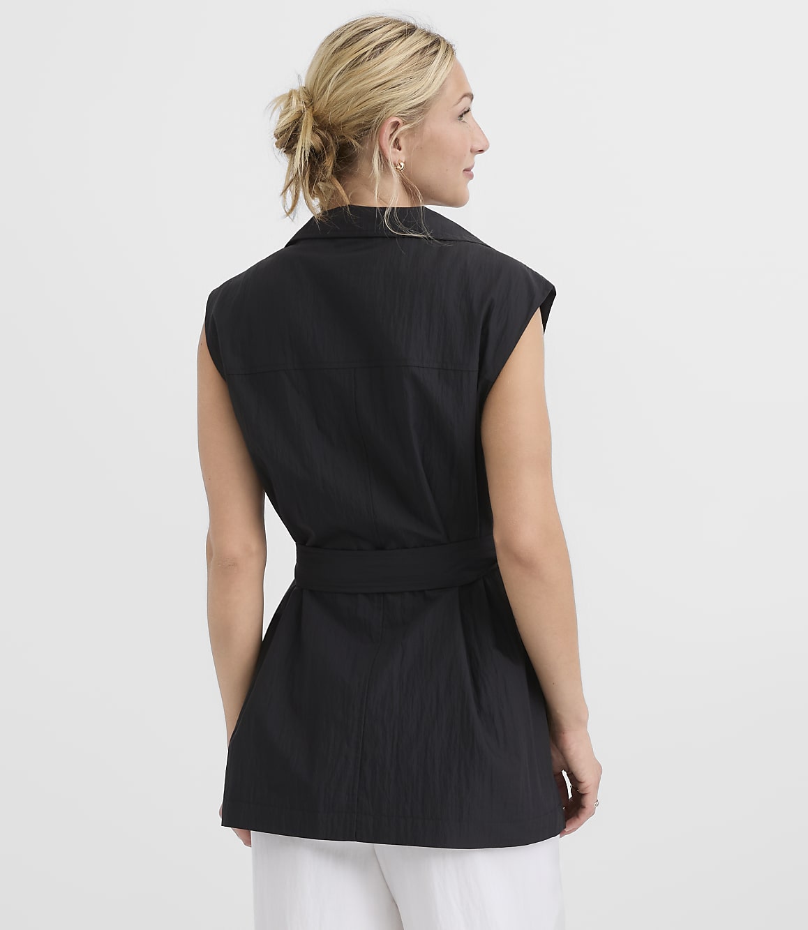 A woman in a black sleeveless belted vest stands against a plain white background.