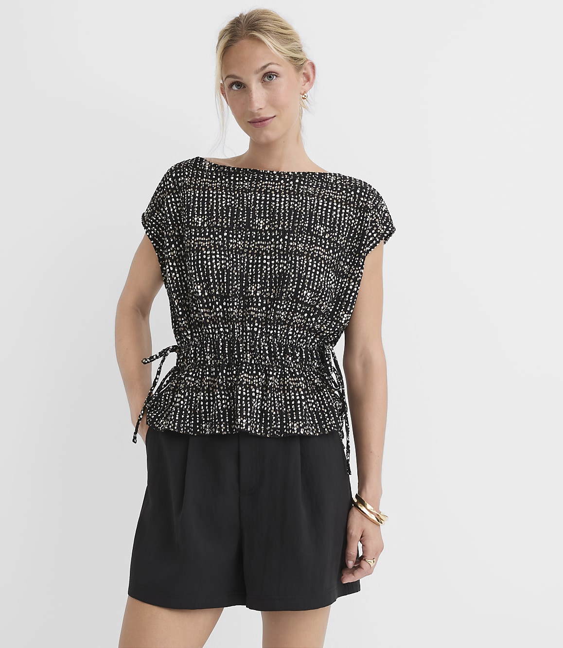 A woman in a patterned top and black shorts stands against a plain white background.