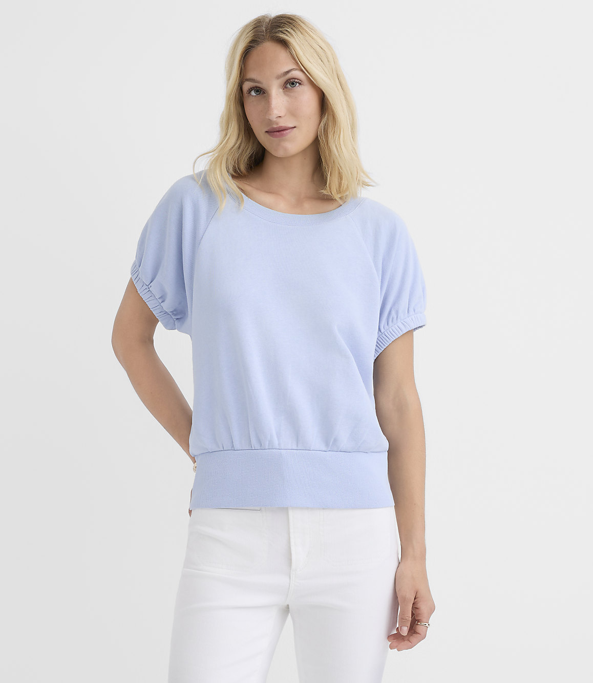 A blonde woman in a light blue top and white pants stands against a plain white background.