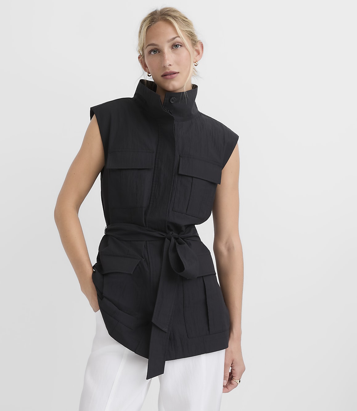 A woman in a black utility vest and white pants stands against a plain white background.