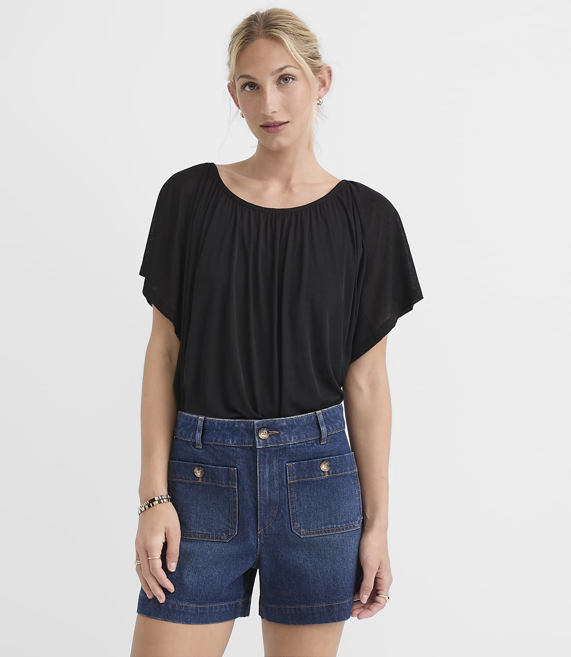 A woman in a black top and denim shorts stands against a plain white background.