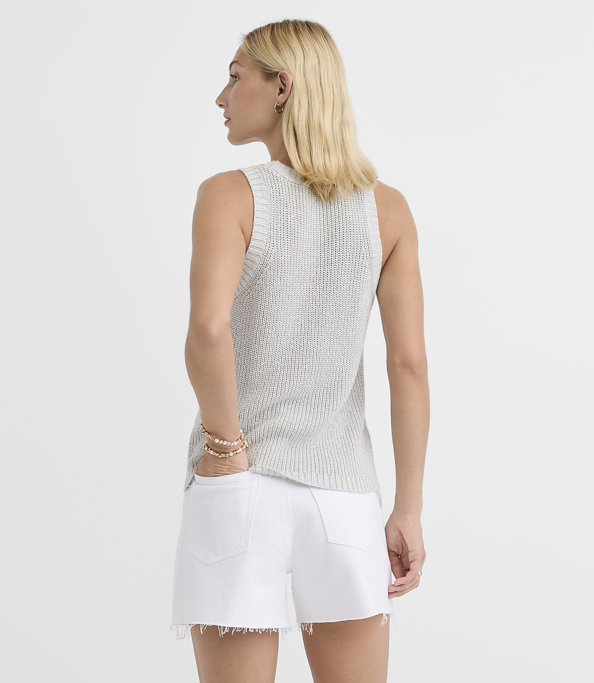 A woman in a light gray knit tank top and white denim shorts stands with her back to the camera against a plain white background.