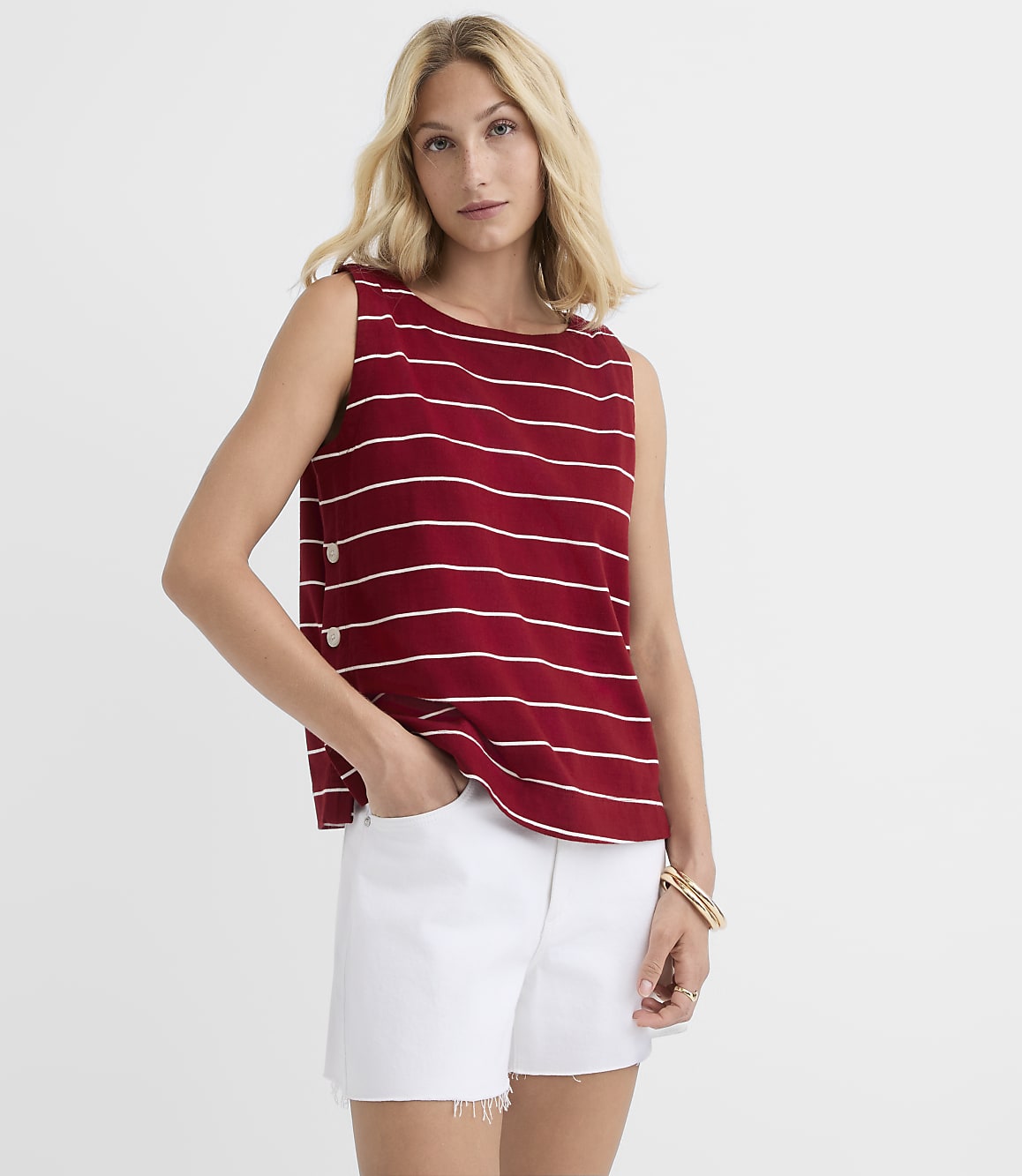 A woman in a red and white striped sleeveless top and white denim shorts stands against a plain white background.