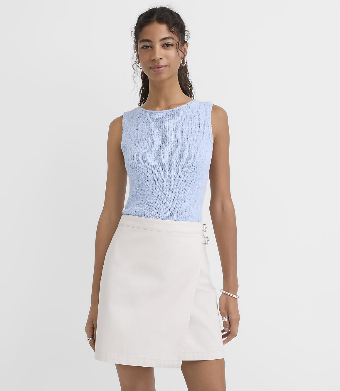 A woman in a light blue textured top and a cream wrap skirt stands against a plain light blue background.