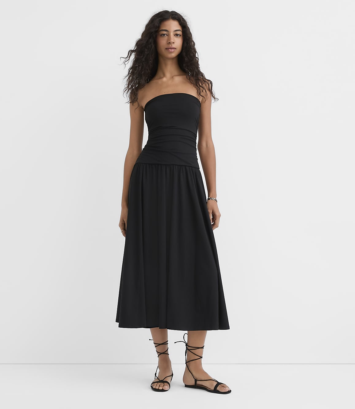 A woman in a black strapless dress and sandals stands against a plain white background.