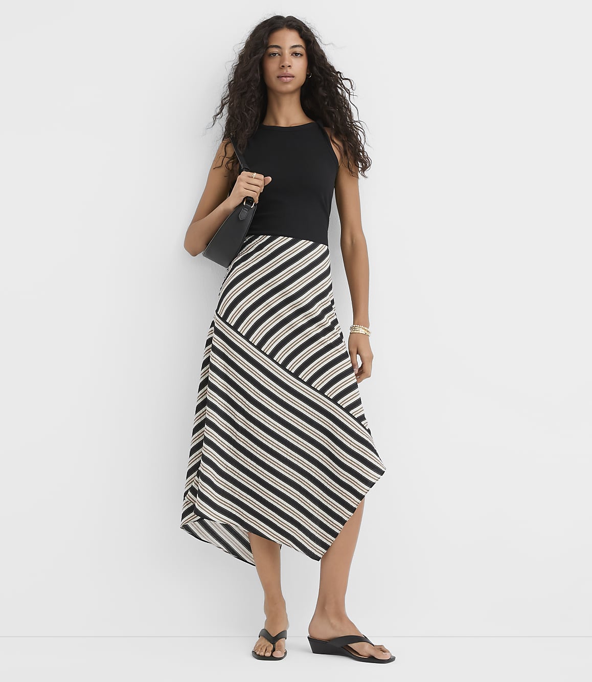 A woman in a black top and a striped asymmetrical skirt stands against a plain white background, carrying a black purse.