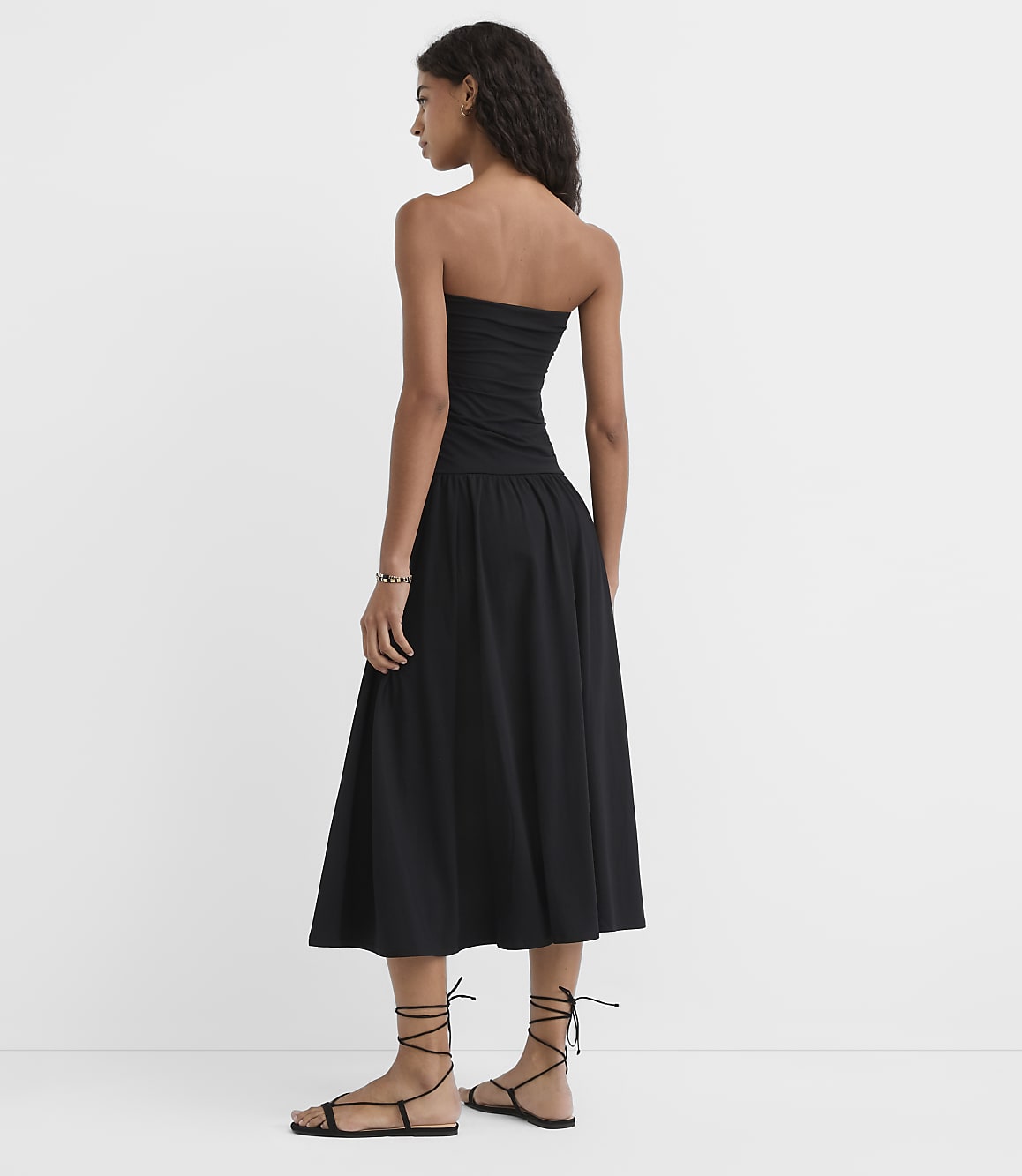 A woman in a black strapless dress and gladiator sandals stands against a plain white background.