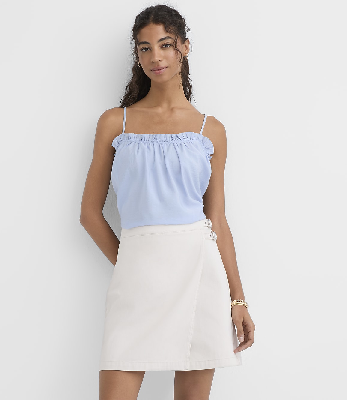 A woman in a light blue ruffled camisole and a cream-colored wrap skirt stands against a plain white background.