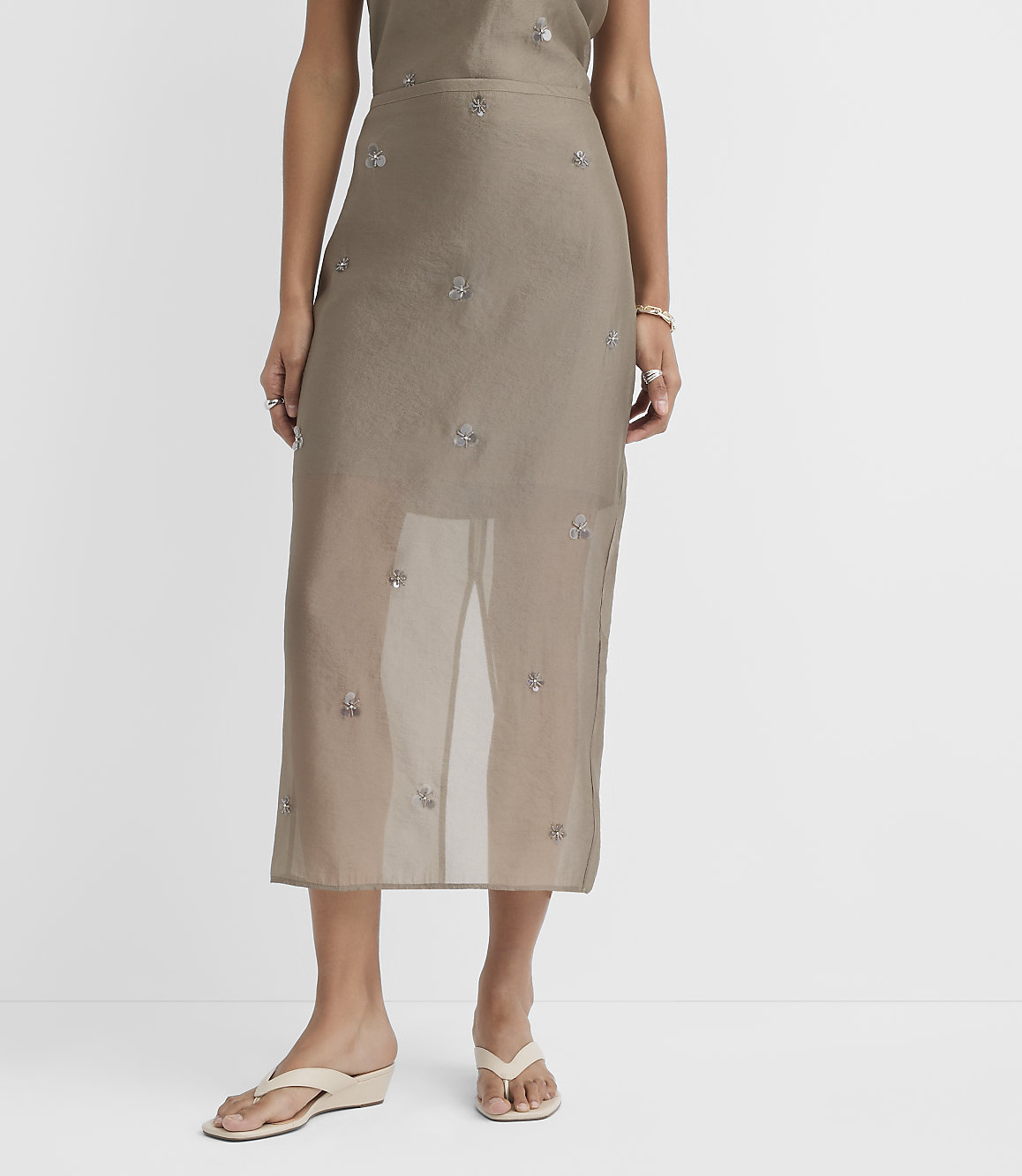 A person stands in a sheer, embellished skirt and cream wedge sandals against a plain white background.
