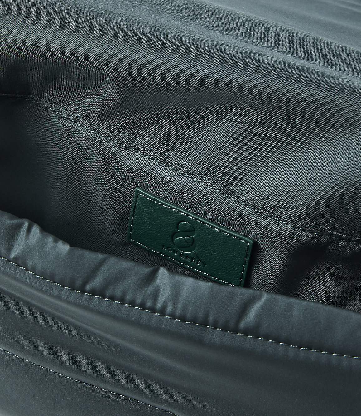 A close-up of a dark green fabric bag's interior reveals a small, embossed green leather tag with a logo in the foreground, against the softly creased, shiny fabric of the bag's lining in the background.