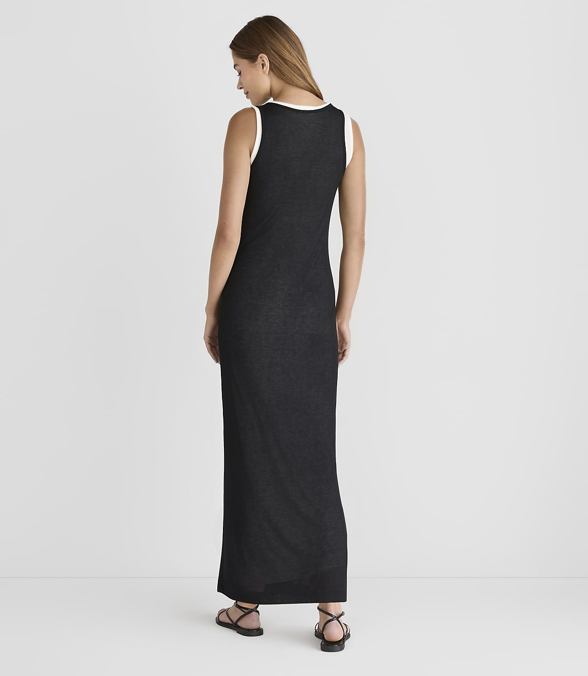 A woman in a black maxi dress with a white trim stands against a plain gray background.